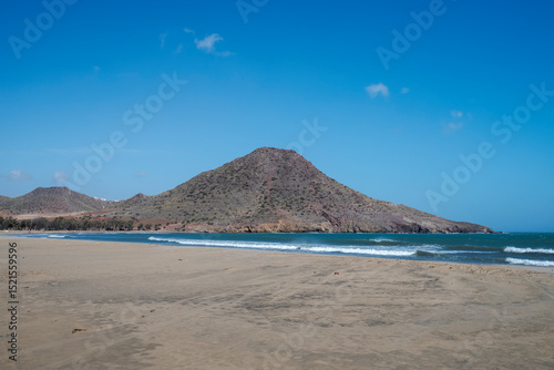 A view of Playa de los Genoveses, Almeria, Spain, with its golden sand, turquoise waters, and a hill in the background under a clear blue sky. A peaceful, unspoiled natural landscape