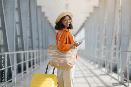 Stylish Black woman with straw bag and yellow suitcase glances over shoulder, happy african american female standing in bright airport hall with phone in hand, smiling at camera, copy space
