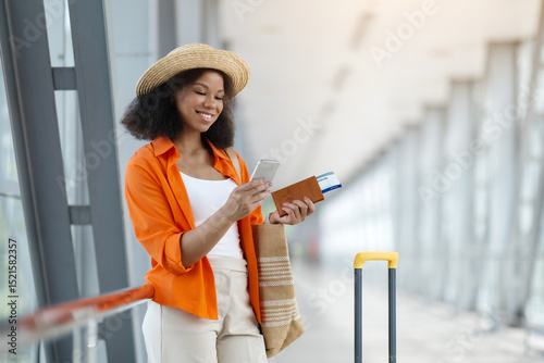 Black woman browsing on cellphone while waiting at airport hall, smiling african american female using travel app or checking flight information in application, navigating her journey in gadget