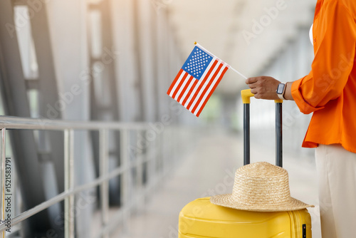 Black female traveler holding American flag and standing by yellow suitcase with summer hat in airport terminal, unrecognizable african american woman ready for trip, cropped