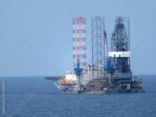 A large offshore oil rig, composed of multiple metal structures, floats on a calm blue ocean under a clear sky. The rig is the central focus, with the ocean forming a solid background. 
