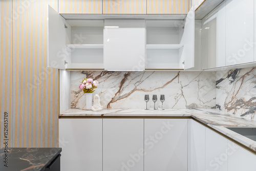 large corner built-in kitchen in white and beige shades. Modern kitchen with upper and lower cabinets and glass display case. white marble countertop and backsplash. Built-in kitchen appliances.