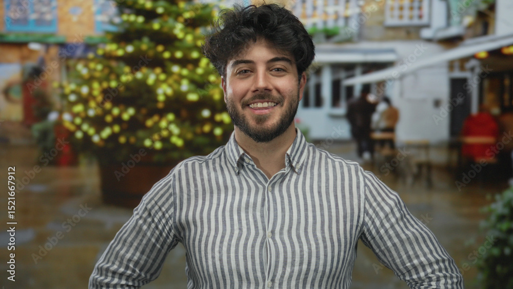 Obraz premium Young hispanic man with beard smiling outdoors on a street with festive lights and a christmas tree, wearing a striped shirt and casual attire, enjoying the holiday atmosphere.