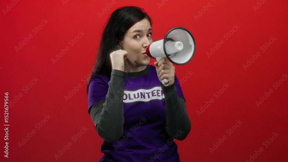 Woman passionately shouting through megaphone wearing volunteer shirt against striking red background with clenched fist for emphasis in an energetic display. Woman passionately shouting through megaphone wearing volunteer shirt against striking red background with clenched fist for emphasis in an energetic display.
