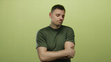 Young man with beard and tattoos stands isolated against a yellow background expressing indecision and contemplation while wearing a casual green shirt.