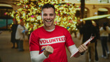 Man in red volunteer shirt smiles holding clipboard with holiday lights in urban area street in background showing cheerful celebration.