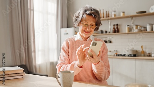 Good-looking mature Caucasian woman with casual clothes spending time in cosy kitchen. Happy grandmother watching positive video or family photo on cell phone. On table standing cup or hot drink.