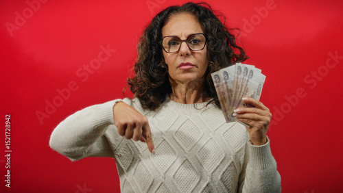 фотографія Woman in glasses pointing down while holding russian rubles against a vibrant red background, appearing serious and focused, wearing a knitted sweater