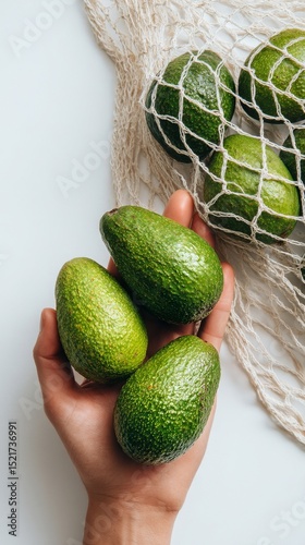 Hand Holding Fresh Green Avocados Beside Mesh Bag on White Fabric.