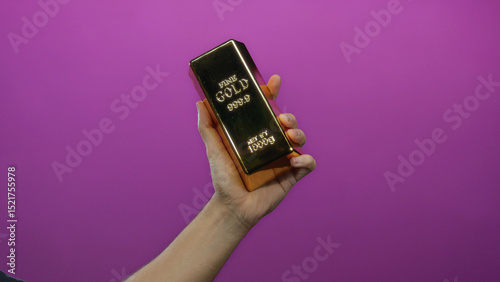 Man holding a fake gold bar against a vivid purple background, showcasing wealth concept in an isolated setting with focus on the hand's firm grip and reflective surface.