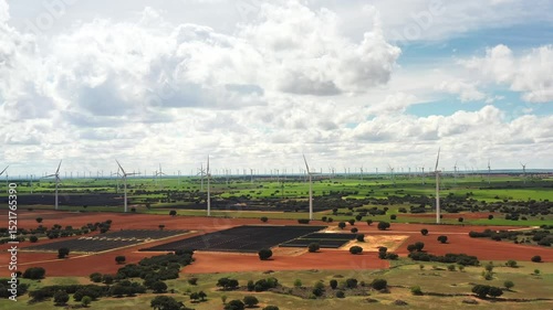 Aerial footage of vast solar and wind energy farm stretching across green and red farmland under bright cloudy sky symbolizing clean energy, sustainability, and environmental innovation