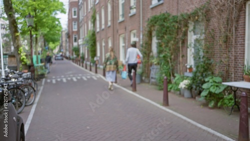 Wallpaper Mural Defocused view of a european city street with a man and woman walking along a brick path lined with greenery, showcasing urban life in a tranquil town setting. Torontodigital.ca