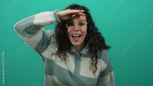 Woman smiling and looking ahead over green background, wearing glasses and striped sweater, young hispanic female standing against isolated vibrant wall.