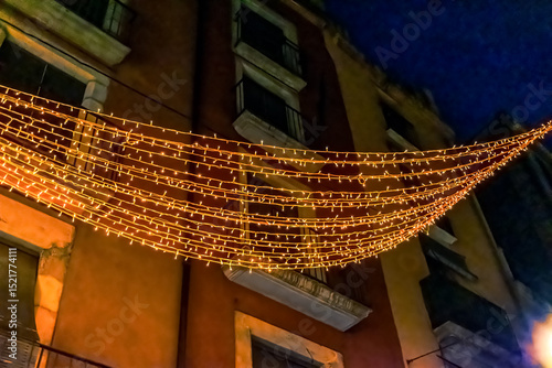 Christmas lights decoration on historic building facade in Girona, Spain. Rows of warm LED string lights creating illuminated canopy against night sky and traditional architecture background