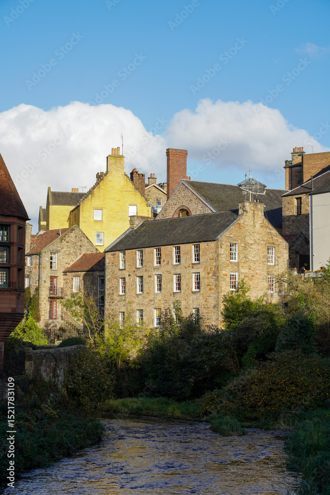 Naklejka premium A sunny afternoon in Edinburgh’s Dean Village, where charming stone houses bask in the golden light, framed by a clear blue sky and fluffy white clouds above the peaceful riverside setting.