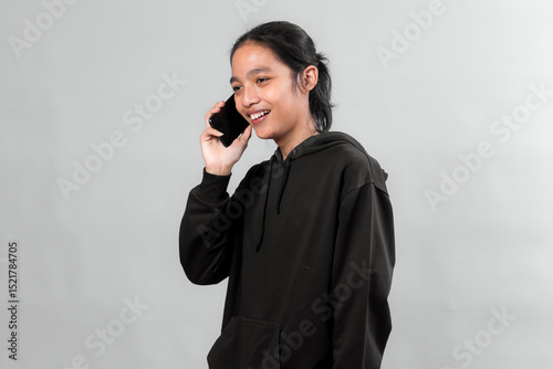 Young Asian man with long hair and black hoodie smiling while talking on a smartphone. Studio portrait against a plain gray background, showing modern lifestyle and communication.
