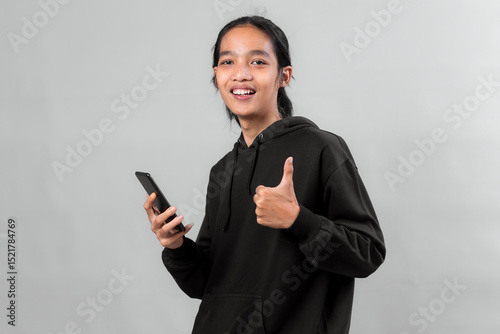 Young Asian man wearing a black hoodie holding a smartphone and giving a thumbs up while smiling. Studio portrait on gray background showing approval, confidence, and modern lifestyle.