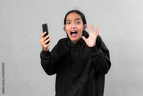 Asian man in black hoodie screaming in shock while holding smartphone, expressing intense fear or panic, isolated on gray background, concept of bad news online.