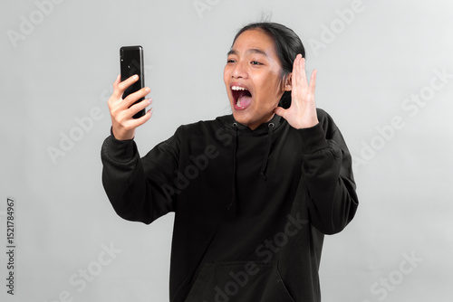 Asian man wearing a black hoodie looks excited and surprised while holding a smartphone during a video call, showing expressive emotion and joyful reaction, isolated on grey background.