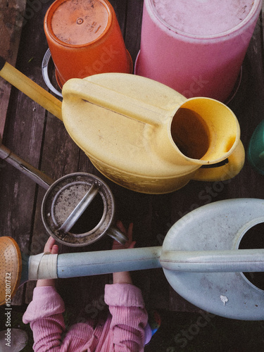 Colorful Gardening Tools and Child's Hands on Wooden Deck – Top View. Rustic gardening scene featuring colorful plastic buckets and watering cans arranged on an aged wooden deck.