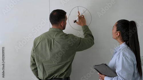 Businessman drawing pie chart on whiteboard, businesswoman watching him with tablet in hand