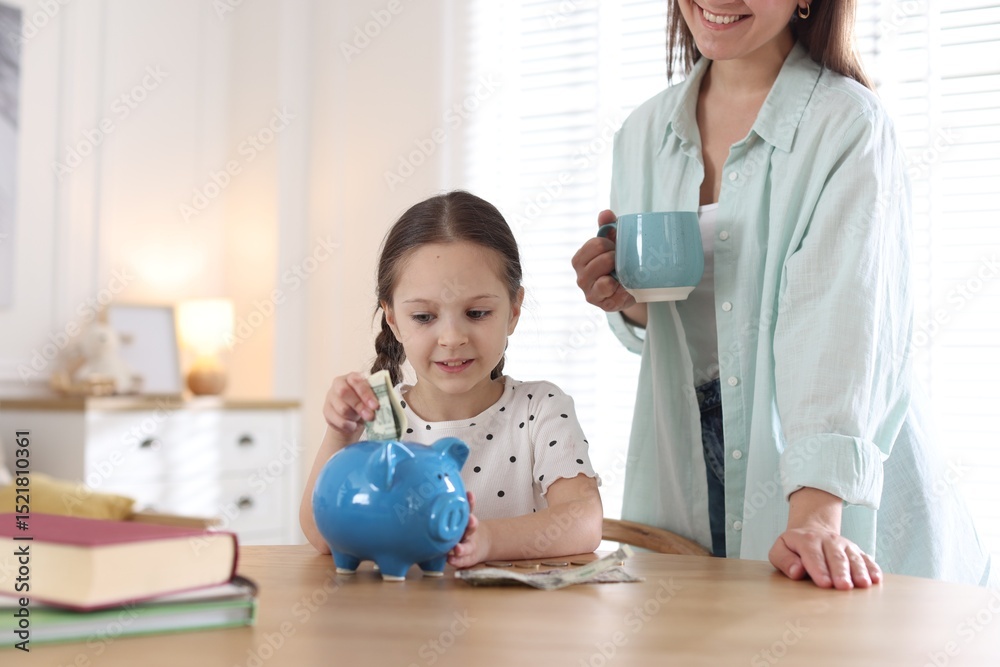 Fototapeta premium Mother and daughter putting pocket money into piggybank at table indoors, closeup