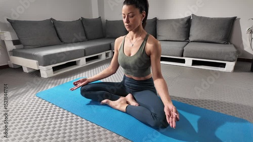 A focused woman engages in mindfulness meditation, seated comfortably on a blue yoga mat in a well-decorated indoor environment. The cozy space features neutral colors and comfortable seating.