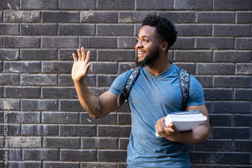 A young man waves cheerfully, holding books, bringing a sense of cheer and openness to his surroundings.