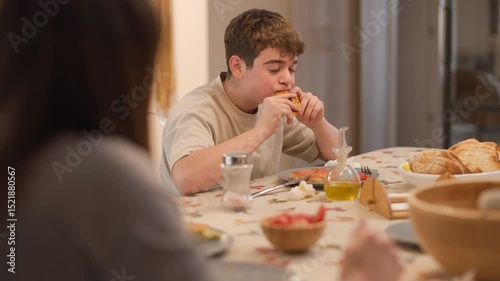 Medium shot of a boy reaching for an empanada from the central plate on the kitchen table during a family meal. The mother stands in the background. Homemade food, relaxed and hungry moment.Horizontal