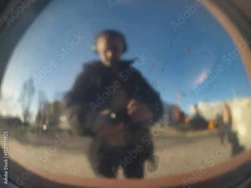 Professional photographer creating distorted self-portrait in curved mirror, holding camera amid contemporary urban architectural setting with dynamic cityscape backdrop