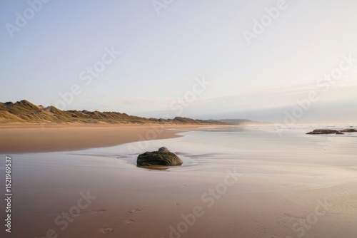Calm expansive beach with soft waves, scattered rocks, distant sand dunes, and gentle morning light creating peaceful atmosphere