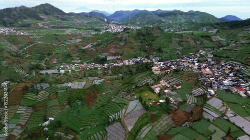 This aerial footage showcases a valley with terraced rice fields, a small village with houses, and distant mountains under a cloudy sky. The camera slowly pans to the left.