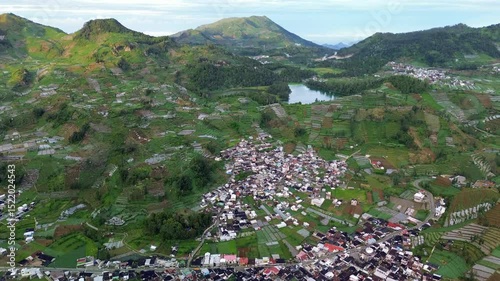 This footage captures an aerial shot of a village nestled among green rice terraces, with mountains and a lake in the background. The camera slowly pans across the landscape.
