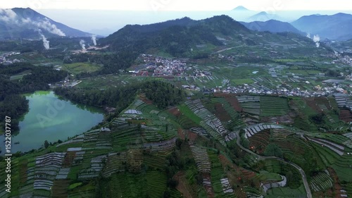 This aerial footage showcases terraced farms, a lake with greenish water, and a village nestled in the Dieng Plateau, Central Java, Indonesia. Mountains visible in the background.