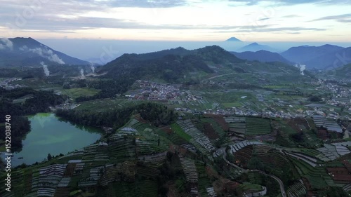 Drone footage captures an aerial view of the Dieng Plateau in Indonesia. Features a lake, terraced agricultural fields, a small town, and distant mountains under a hazy sky.