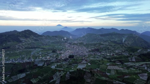 This aerial footage showcases a town nestled in a valley, surrounded by lush green terraced fields. Mountains rise in the background under a soft sunrise sky.