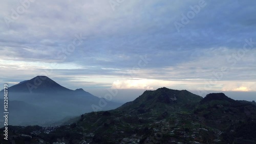 A wide shot of a mountain range under a cloudy sky. The sun is rising or setting, casting a soft light on the landscape. There is a small town in the valley.