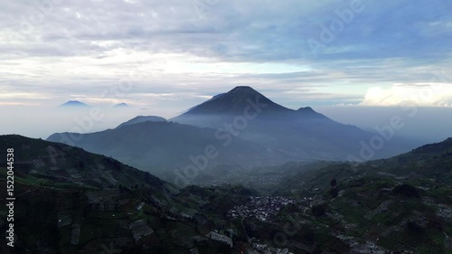 Aerial footage captures a mountain range with a village nestled below. Terraced fields cover the surrounding hillsides under a cloudy sky. Camera is static