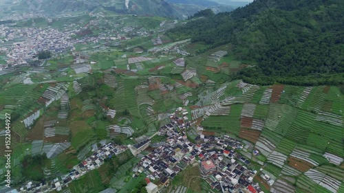 This aerial footage showcases a village nestled among lush, green terraced rice fields in the Dieng Plateau. The camera slowly pans across the landscape, revealing the intricate patterns of the fields