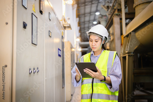 Engineer wearing safety helmet and vest inspects electric grid switchgear in industrial field using tablet to ensure gas insulated system operates safely and efficiently