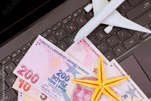 Fototapeta Naklejka Na Ścianę i Meble -  Currency from Turkey lira banknotes lies on laptop keyboard alongside travel plane starfish, symbolizing vacation planning.