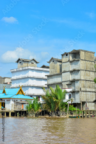 A riverine settlement in a tropical location, a mix of traditional stilt houses and more industrial-looking buildings, situated along a murky brown river. Can represent urban development in river life