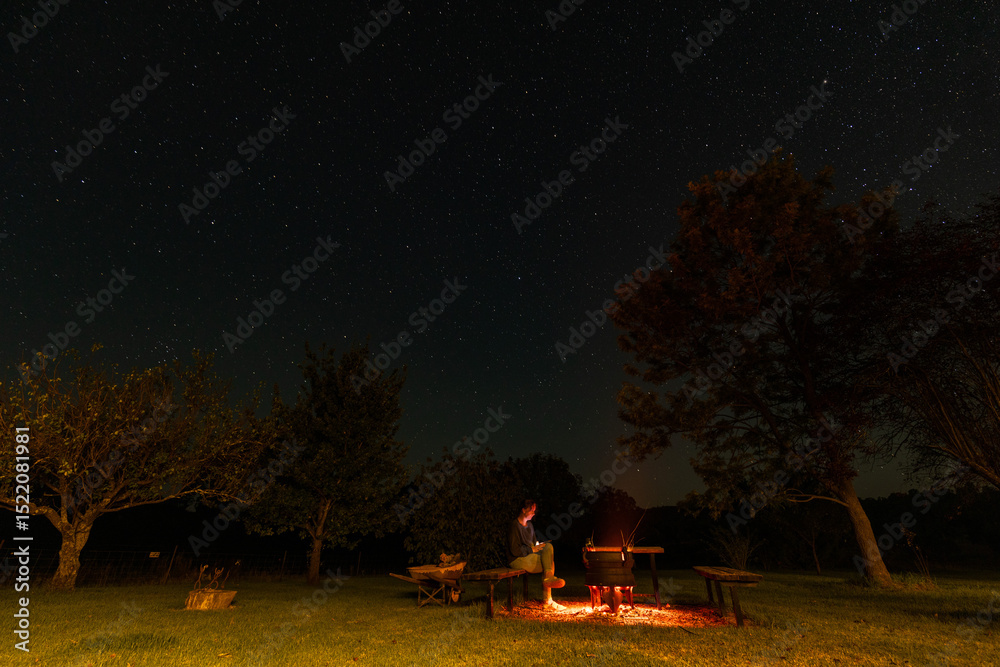 Fototapeta premium Woman sitting by backyard fire pit looking up at the stars in the night sky above