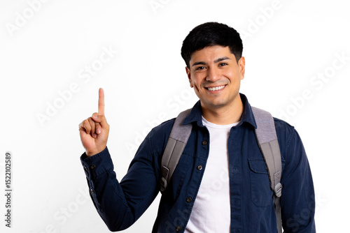 Young latin man, carrying backpack, looking at camera, pointing to a copy space on white background