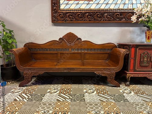The image showcases a beautifully carved wooden daybed or settee with intricate details and a rich finish, positioned on a patterned rug in what appears to be an elegantly decorated living space, flan
