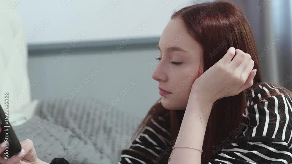 Teenage girl with long red hair relaxing on bed, casually using smartphone in cozy bedroom, engaging with digital world through mobile device