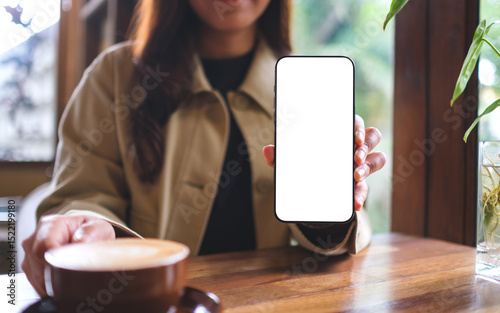 Mockup image of a woman holding and showing mobile phone with blank white screen in cafe