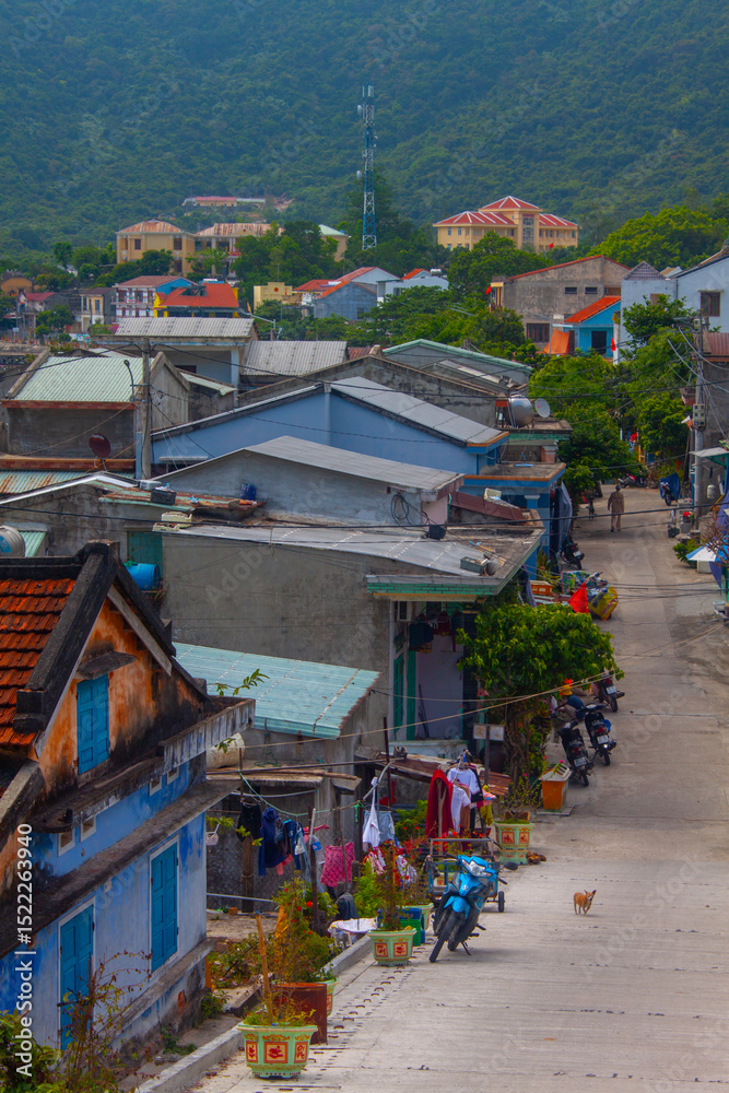Fototapeta premium Colorful Vietnamese village street with traditional houses, scooters, laundry hanging, and mountains in the background. A small dog walks along the quiet road. 