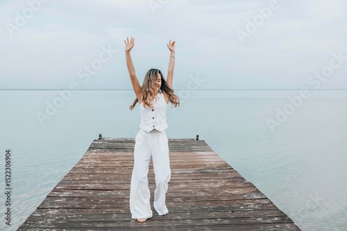 woman dancing barefoot on wooden pier, long hair flowing as she twirls freely under a pale sky, surrounded by calm water, joyful expression in motion, freedom, wellness, motion blur