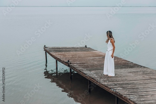 unrecognizable woman in white outfit stands on wooden pier, calm lake, peaceful overcast, viewed from behind with contemplative mood, mindfulness, travel retreats, wellness